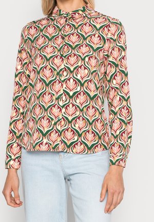 Kvinde iført en knaplukket langærmet skjorte med et pink, guld og grønt blomsterprint samt lyseblå jeans mod en ensfarvet baggrund.