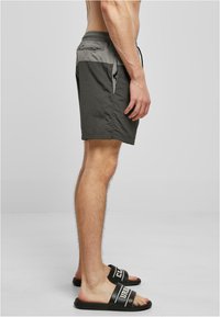 Graue Badeshorts mit Kordelzugbund, Seitentaschen und einer hinteren Reißverschlusstasche. Getragen mit schwarzen Slide-Sandalen mit einem Logoband.
