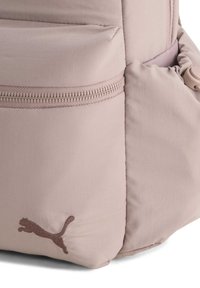 Sac à dos rose clair avec un tissu texturé, une fermeture éclair proéminente et un logo brodé marron représentant un chat sautant sur le côté.