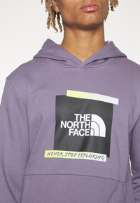 Sudadera morada hecha de una tela suave con un parche de logo negro y una banda de acento colorida que dice "NUNCA DEJES DE EXPLORAR."