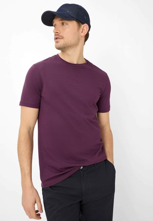 Mann trägt ein lila kurzärmeliges Hemd, schwarze Hose und dunkelblauen Schirmmütze, schaut nach links vor einem weißen Hintergrund.