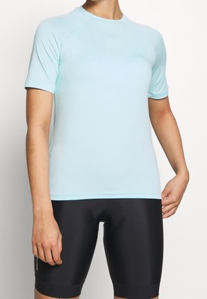 Person iført en lyseblå kortærmet sportsbluse og sorte polstrede cykelshorts mod en ensfarvet baggrund.