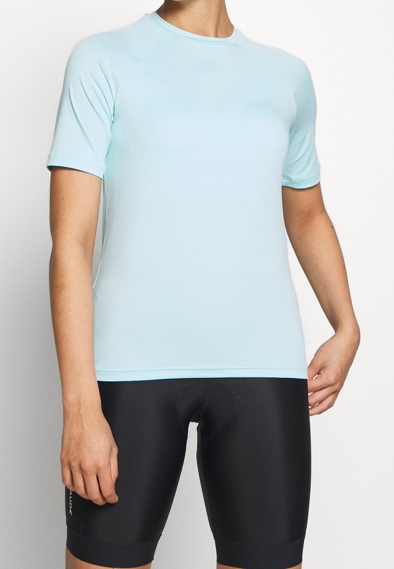 Personne portant un maillot de sport bleu clair à manches courtes et un cuissard de cyclisme rembourré noir sur un fond uni.
