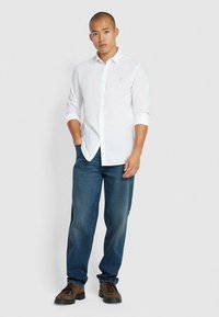 Camicia bianca con maniche arrotolate, abbinata a jeans blu scuro e scarpe marroni. La camicia presenta un piccolo logo sul petto.