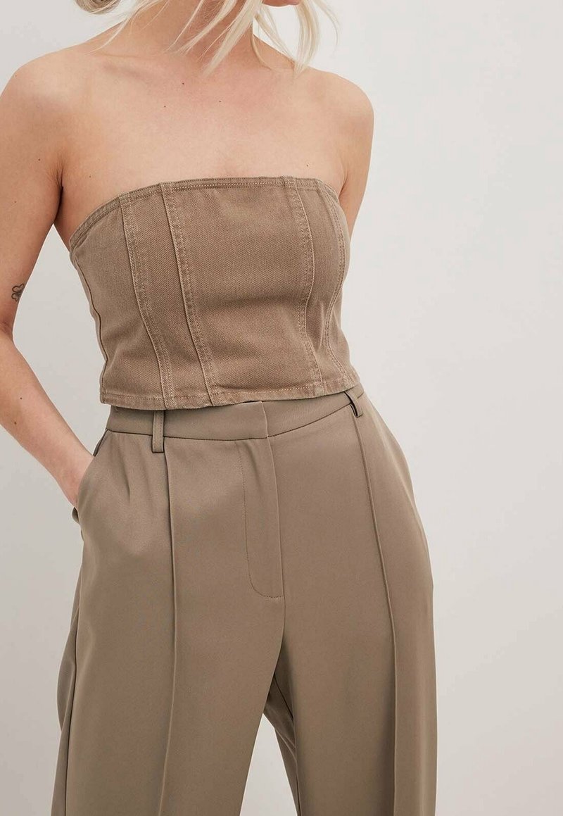 Femme portant un haut style corset marron sans bretelles et un pantalon beige taille haute à jambes larges, les mains dans les poches, sur un fond uni.