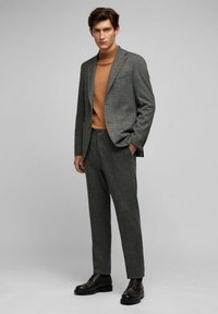 Grau karierter Anzug mit taillierter Passform, kombiniert mit einem sandfarbenen Rollkragenpullover. Schwarze Lederschuhe, glatte Textur und klare Linien im gesamten Outfit.