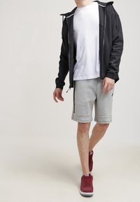 Man i svart zip-up huvtröja, vit t-shirt, grå shorts och röda gympaskor, som går mot en plain ljus bakgrund.