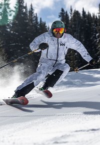 L'équipement de ski comprend une veste à motifs blancs et gris avec des accents noirs, associée à un pantalon noir et des chaussures de ski distinctes rouges et noires.