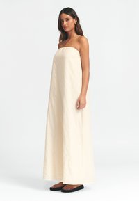 Brezramna maksi obleka iz mehkega kremnega blaga s subtilnimi teksturiranimi črtami. Ohlapen kroj, tekoča oblika, v kombinaciji z ravnimi sandalami.