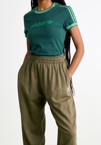 Žena nosí zelené tričko Adidas s logem a pruhovanými rukávy, doplněné olivově zelenými sportovními kalhotami Adidas s elastickým pasem a kapsami.