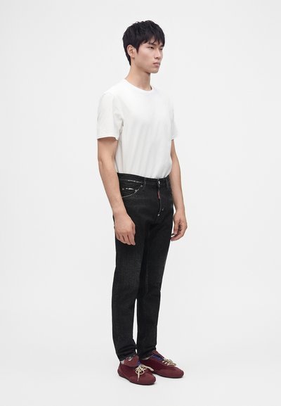 Homme debout en t-shirt blanc, jean noir et baskets rouges sur un fond blanc uni, légèrement tourné vers la droite.