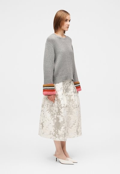 Pilkas megztinis su spalvotomis dryžuotomis rankogaliais, derinamas su baltu, kelio ilgio gėlių rašto sijonu ir baltais kulniukais su atvirais pirštais.