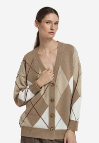 Beżowy kardigan w wzór argyle z brązowymi, białymi i czerwonymi diamentami. Miękki materiał dzianinowy, dekolt w serek, zapięcie na guziki i ściągane mankiety.