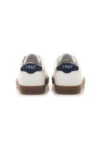 Baskets en cuir blanc avec un empiècement de talon bleu marine arborant "1967" en texte blanc. La semelle est marron clair et le design est bas.