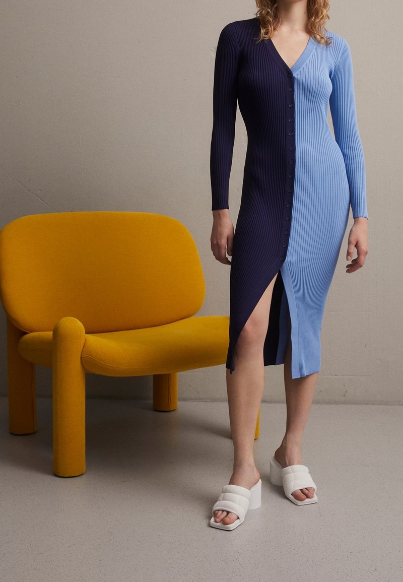 Robe côtelée à manches longues, présentant un design à découpe en bleu marine foncé et bleu clair, complétée par des sandales à talons bloc blanches et une chaise jaune.