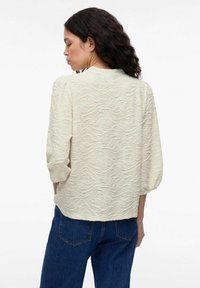 Cream-colored blouse with textured wave pattern, three-quarter sleeves, and a rounded hem, paired with blue jeans.