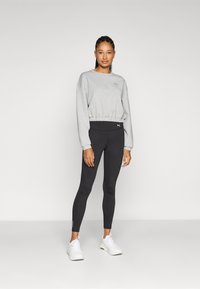 Suéter gris corto con dobladillo elástico, mallas negras ajustadas y zapatillas deportivas blancas, mostrando un logo en la parte superior izquierda.