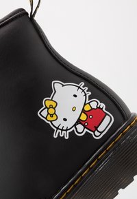 Černá kožená bota s žlutým švem a bílou kreslenou kočkou v červeném oblečku a žlutou mašlí po boku.