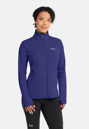 Vrouw met kort krullend haar, gekleed in een donkerblauw jasje met rits en een zwarte broek, staande tegen een eenvoudige lichte achtergrond.