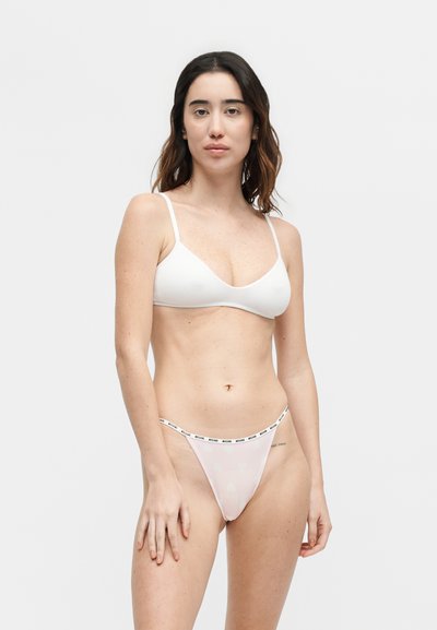 Femme aux longs cheveux foncés portant un bralette blanc et un string rose clair avec une ceinture à logo, debout devant un fond blanc uni.