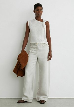 Kobieta w białym topie bez rękawów i szerokich białych spodniach, trzymająca brązową kurtkę, stojąca na tle gładkiej białej ściany i szarej podłogi.