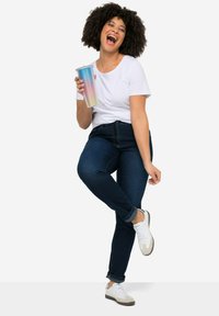 Biela tričko s krátkym rukávom, tmavomodré tesné džínsy a biele tenisky. Drží farebný textúrovaný pohár s viečkom a slamkou.