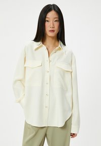 Camicia button-up beige chiaro con colletto, due tasche sul petto e maniche lunghe. Tessuto liscio, vestibilità casual e orlo curvo.