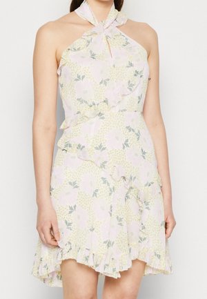 Ærmeløs kjole med halterneck og blomsterprint, med flæser, i pastelpink og gul, båret af en person stående mod en ensfarvet baggrund.