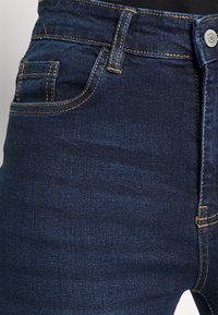 Jeans de denim azul escuro com costuras amarelas em contraste, design de cinco bolsos e um botão prateado na cintura. Textura suave.