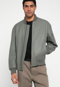 Nahkainen pommitakki hillityssä vihreässä, jossa on vetoketjullinen suljenta, resorit hihansuissa ja sivutaskut. Yhdistetty mustaan paitaan ja beigenvärisiin housuihin.