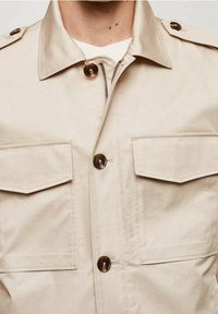 Beżowa kurtka z kołnierzem, dwoma kieszeniami na wysokości klatki piersiowej i zapięciem na guziki z przodu. Wykonana z teksturowanej tkaniny z brązowymi guzikami.