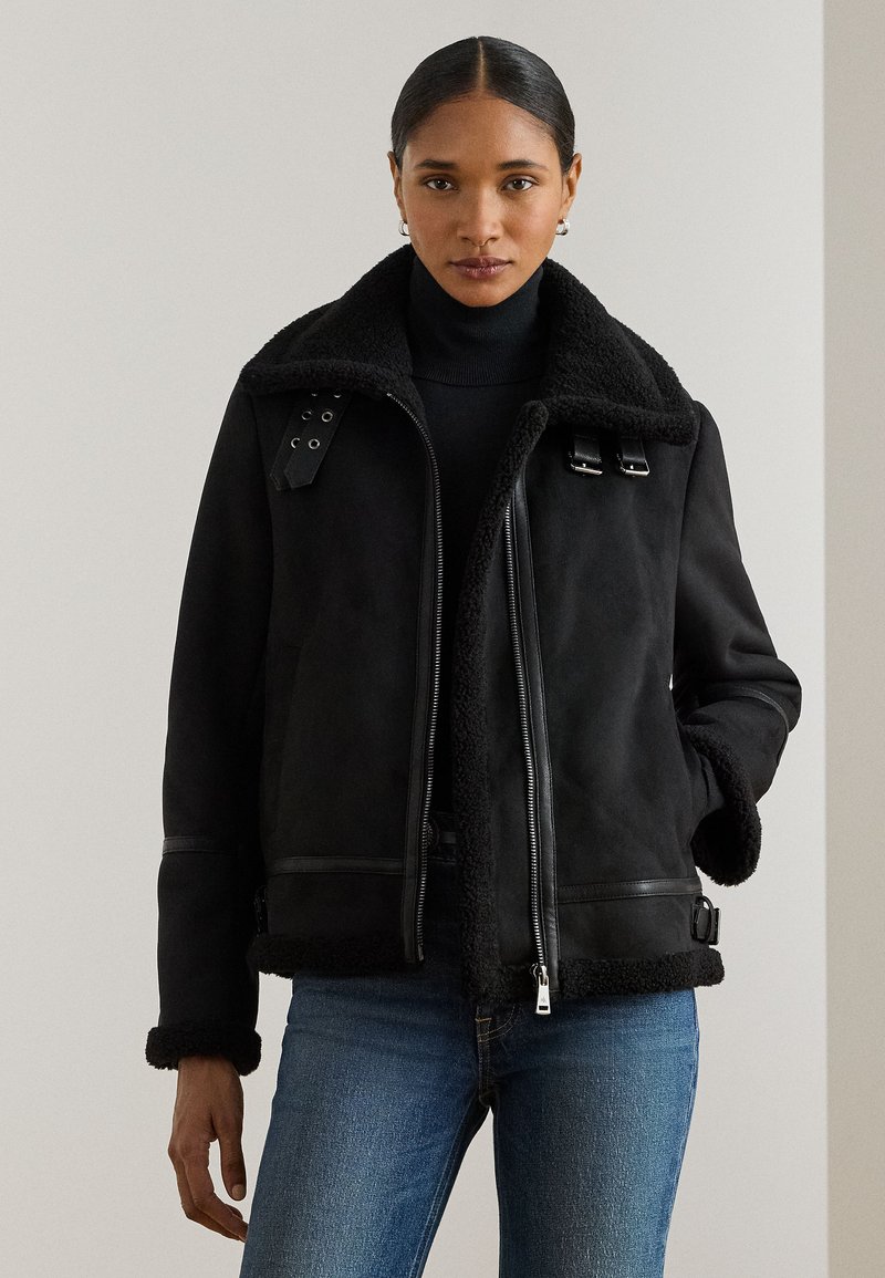 Femme portant une veste noire doublée en peau lainée, un col roulé noir, un jean bleu, cheveux tirés en arrière, une main dans la poche, fond neutre.
