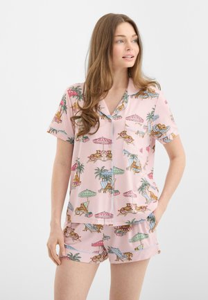 Mujer con pijama de dos piezas de manga corta y pantalones cortos de color rosa con estampados de tigres, descansando bajo sombrillas de playa y palmeras.