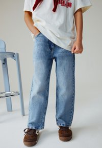 Jeans in denim azzurro chiaro con una vestibilità rilassata, accompagnati da sneakers marroni con motivo a stampa leopardo. Lo sfondo è neutro.