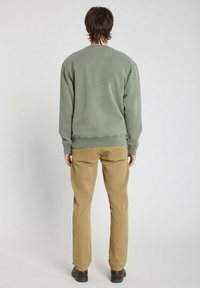 Sweatshirt vert avec des poignets et un ourlet côtelés, associé à un pantalon beige, vue de dos sur un fond gris clair.