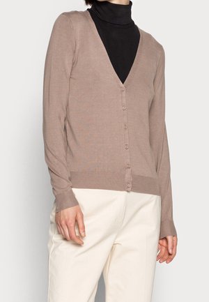 Donna che indossa un cardigan beige con bottoni sopra un dolcevita nero e pantaloni bianchi, in piedi davanti a uno sfondo chiaro e uniforme.