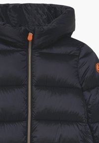 Marineblauwe gewatteerde jas met hoge kraag, zilveren rits met oranje trekker en een oranje ronde patch op de mouw.