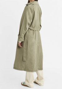 Vaaleanoliivinvihreä trenssitakki puuvillatwillistä, jossa on rento leikkaus, vyö vyötäröllä ja käärityt hihat. Yhdistetty leveisiin kermaisiin housuihin ja kenkiin.