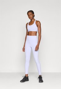 Σετ αθλητικό μπουστάκι και κολάν ανοιχτό μωβ, εφαρμοστή γραμμή, με ομαλή υφή και λογότυπο στο πάνω μέρος. Μαύρα αθλητικά παπούτσια ολοκληρώνουν την εμφάνιση.