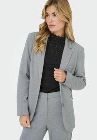 Pilkas pritaikytas švarkas, pagamintas iš minkštos medžiagos, su išpjautu apykakle, priekinėmis kišenėmis, derinamas su juodu nėriniuotu marškinėliu. Modelis pozuoja atsipalaidavęs.