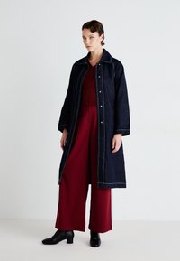 Langer dunkler Denim-Mantel mit heller Naht, kombiniert mit einem burgunderfarbenen Oberteil und weiten Hosen. Das Model trägt schwarze, hochhackige Stiefeletten.