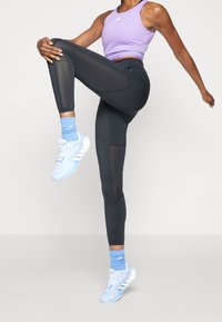 Leggings de sport noir avec des accents en maille, associés à un crop top lavande et des chaussures et chaussettes de sport bleu clair, dans une pose dynamique.