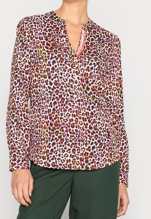 Kobieta ubrana w bluzkę z długim rękawem w panterkę z różowymi, pomarańczowymi i zielonymi akcentami, w połączeniu z ciemnozielonymi spodniami, stojąca nieruchomo.