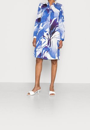 Vrouw draagt een knielange, lange mouwen overhemdjurk met een blauw-wit abstract bladpatroon, gecombineerd met witte sandalen met hak.