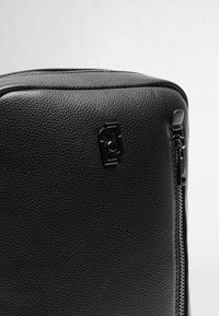 Borsa in pelle nera con una texture a granuli, dotata di chiusura con zip e un dettaglio logo nero sul davanti. Forma rettangolare semplice.