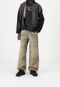 Osoba v tmavě šedé bundě přes černé tričko s potiskem, béžových opotřebovaných cargo kalhotách s kolenními záplatami a černých teniskách, stojící na bílém pozadí.