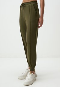 Olijfgroene joggingsbroek met elastische tailleband en trekkoord, voorzien van twee zijzakken en taps toelopende boorden. Draag met witte sneakers.
