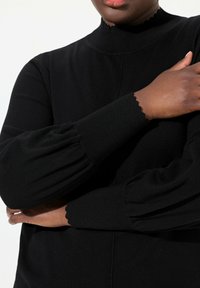 Mustat ribbattu neulepaita, jossa on korkea kaulus ja aallotetut hihansuut, sekä kuplamaiset hihat, jotka antavat tekstuuria. Lähikuva kädestä ja käsivarresta.