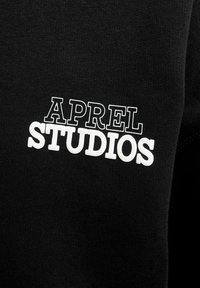 Schwarzer Stoff mit einer glatten Textur, der den Text "APREL STUDIOS" in fetten, weißen Buchstaben zeigt und so einen Kontrast zwischen den Farben schafft.