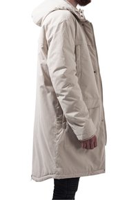 Parka beige avec capuche, dotée d'une fermeture éclair, de deux poches avant et d'un tissu légèrement texturé. Silhouette mi-longue.